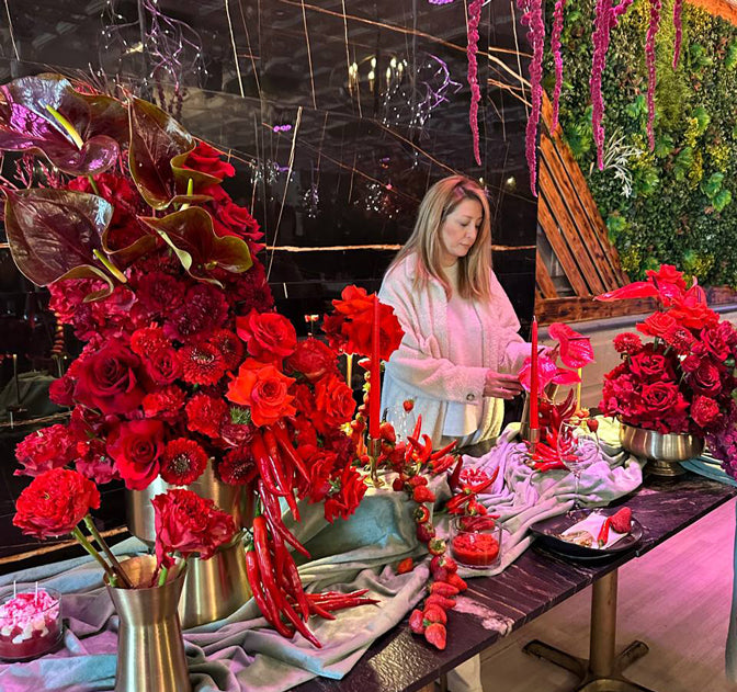 Woman arranging flowers in a vibrant, decoratively rich setting with red floral arrangements and gold vases. Lotus Land Flowers Boutique